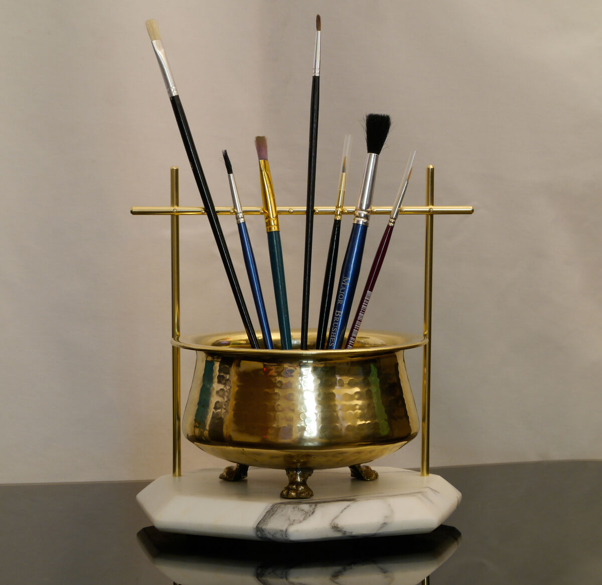 Hammered brass pot on a white marble base, paintbrushes hanging from a brass rail